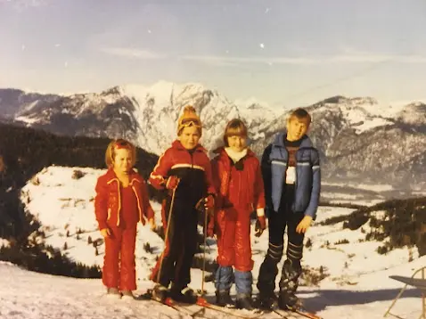 Old photo of four children in red and blue ski clothing with skis in the snow, with mountain peaks behind them
