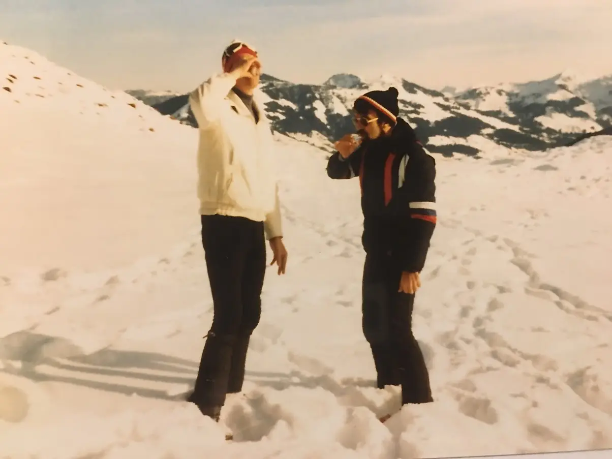 Old photo of two men in ski clothing standing in the snow having a drink, with snowy mountains behind them