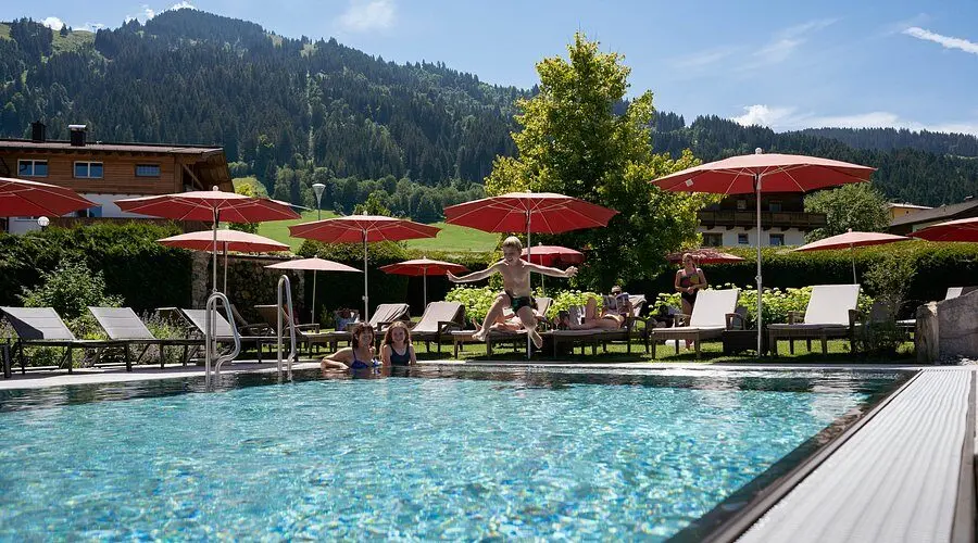 Kind springt in het buitenzwembad van Hotel Wastlhof, met parasols en bergen op de achtergrond