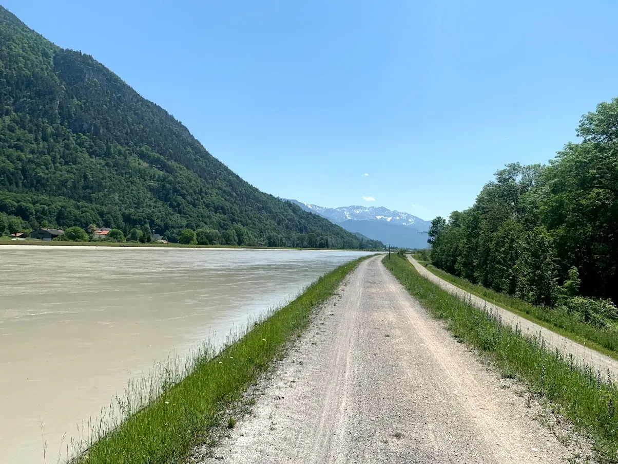 Gravel path along a wide, silty river in a valley with mountains and snowy peaks in the distance