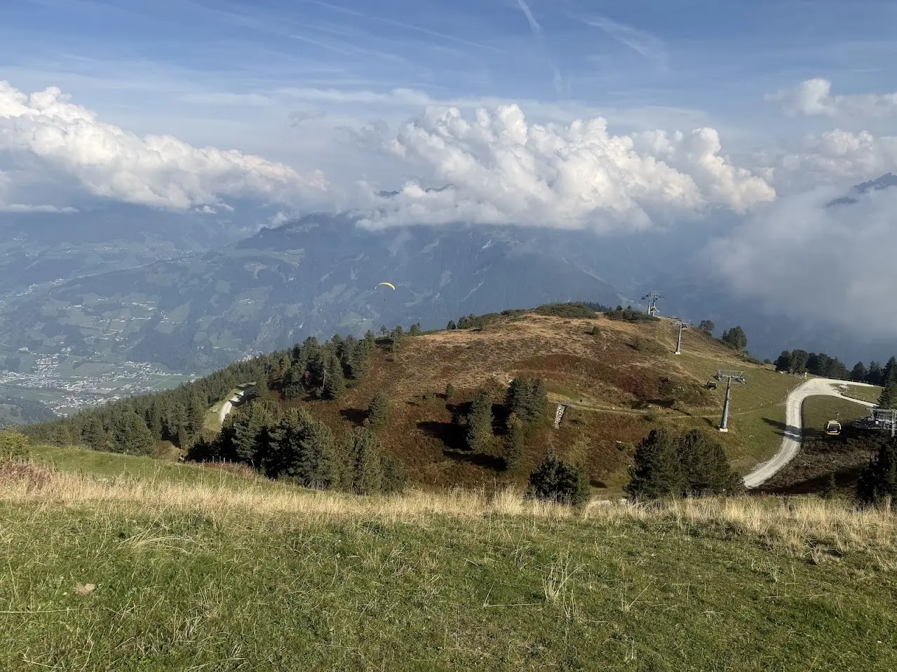 Autumnal mountain plateau with a gondola lift, a yellow paraglider in the distance and a valley with villages beneath clouds