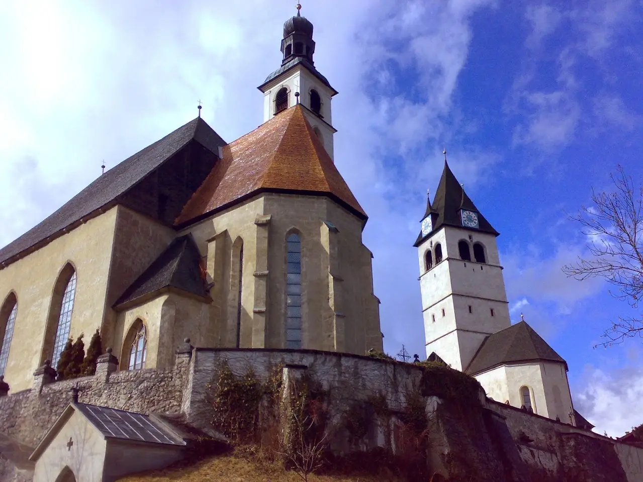 Church with a red roof, a small onion-shaped tower and a second clock tower, above a stone wall under a blue sky with clouds