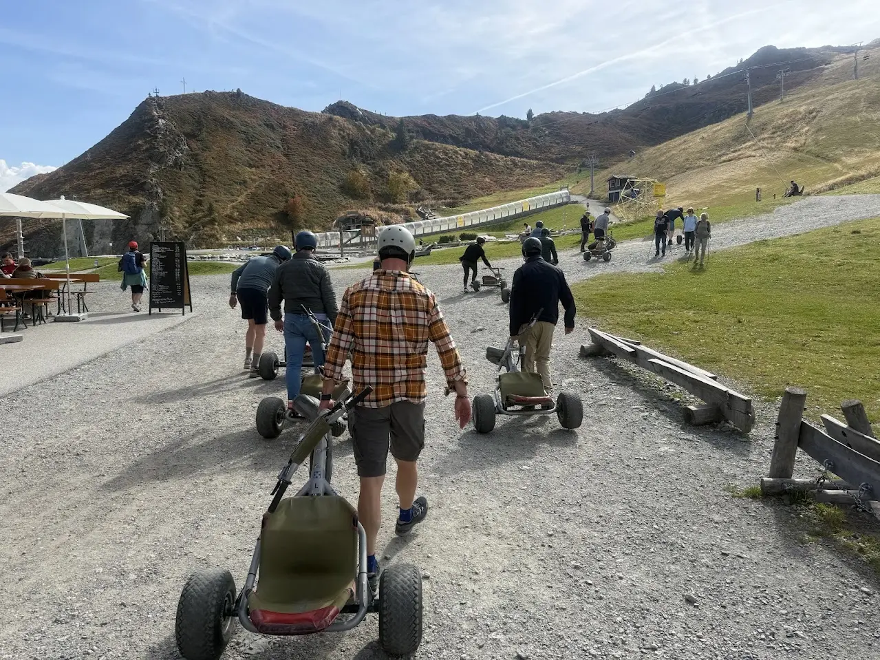 Group of people with helmets pulling mountain carts along a gravel path towards the start of a track, with mountain peaks and a chairlift behind