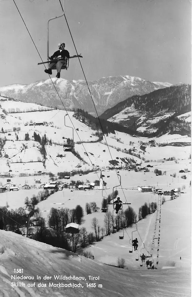 Ancienne carte postale d’un skieur sur un télésiège au-dessus d’une vallée enneigée avec une église et un village, légendée « Skilift auf das Markbachjoch, 1455 m »
