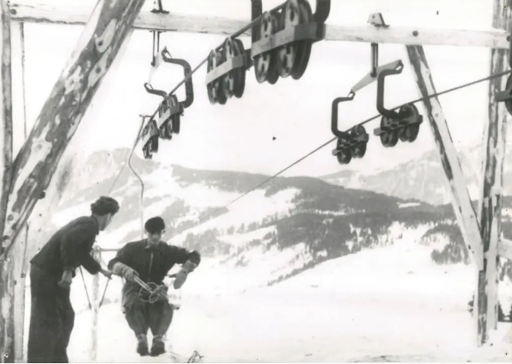 Photo en noir et blanc de deux hommes à côté d’un siège sous les galets d’une station supérieure en bois, avec des collines enneigées derrière
