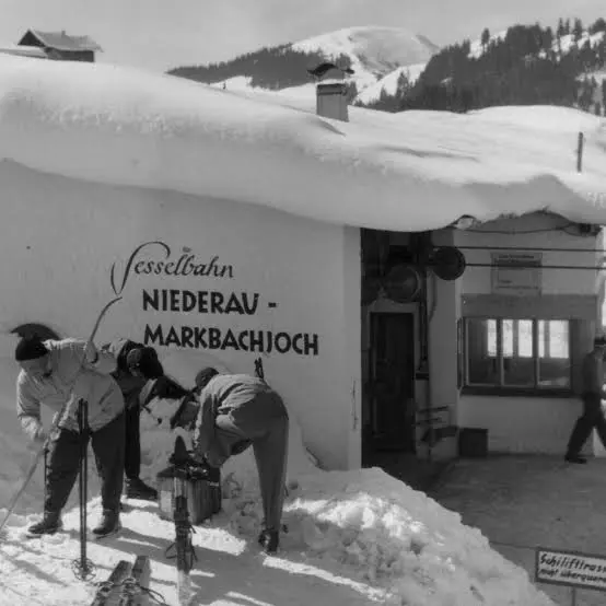 Photo en noir et blanc de skieurs dans la neige préparant leurs skis devant une gare portant l’inscription « Sesselbahn Niederau – Markbachjoch »