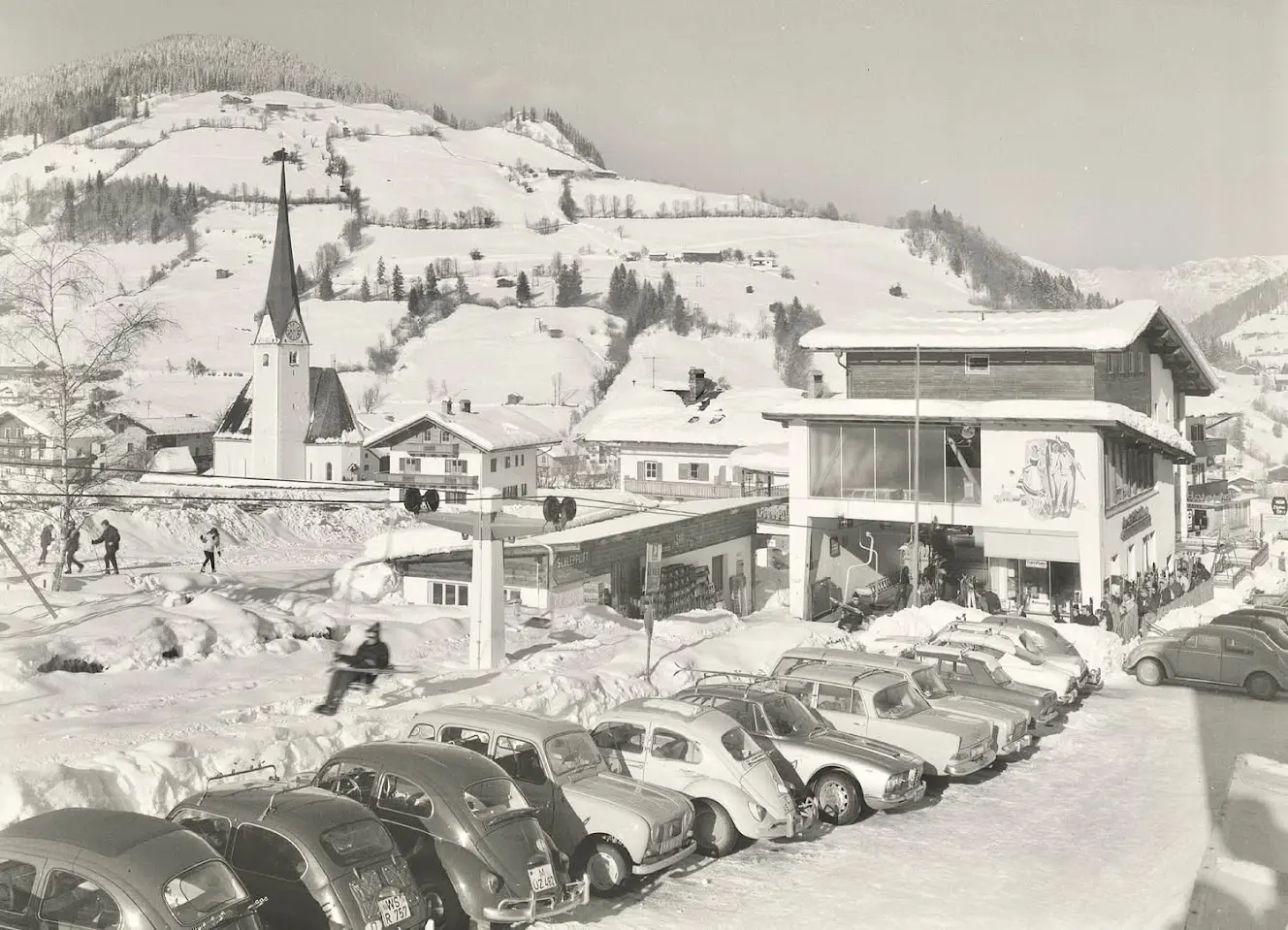 Ancienne photo en noir et blanc du village en hiver avec des voitures anciennes garées, une église et un bâtiment avec une gare de remontée vitrée