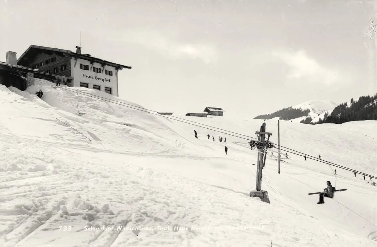 Ancienne photo en noir et blanc d’un télésiège avec des skieurs sur une large piste enneigée, avec la Haus Berglift tout en haut