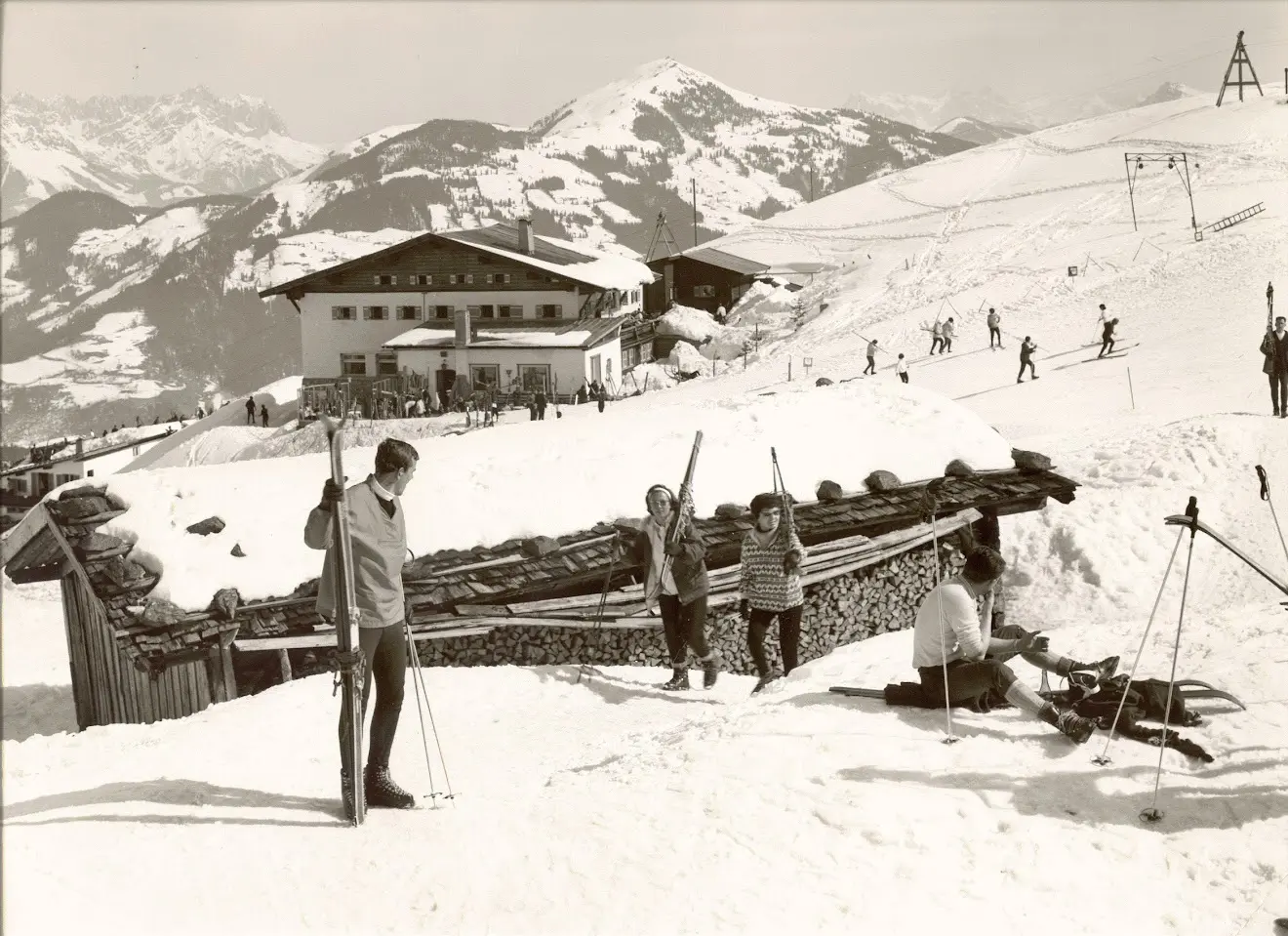 Ancienne photo en noir et blanc de skieurs avec des skis en bois près d’une maison de montagne et d’un abri à bois dans la neige, avec des montagnes et un téléski derrière