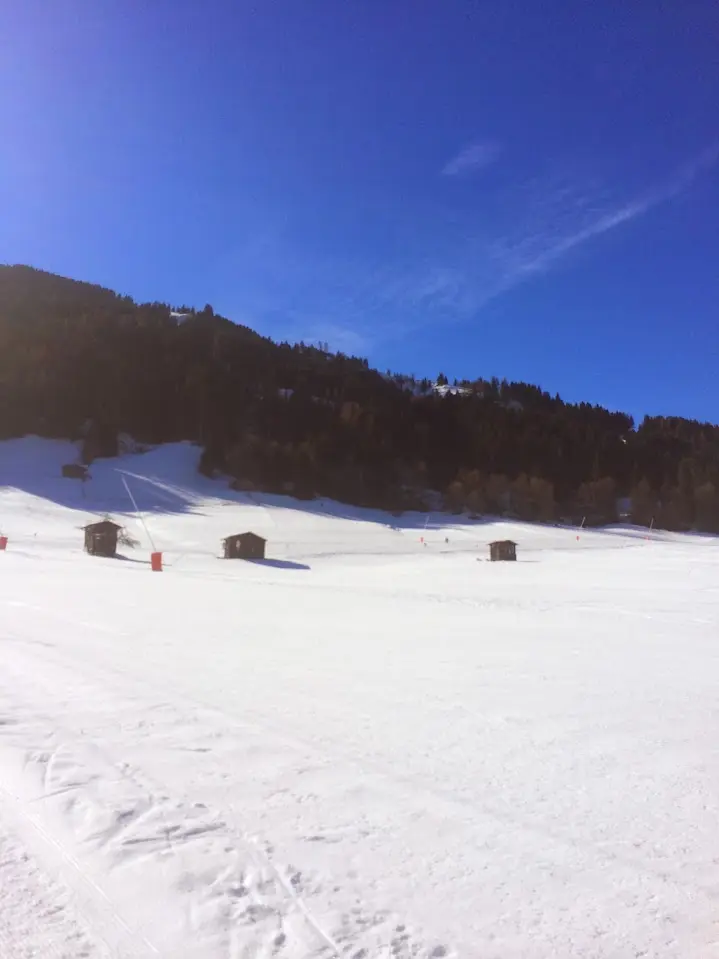 Wide, sunny ski slope with small wooden huts and a snow cannon under a deep blue sky