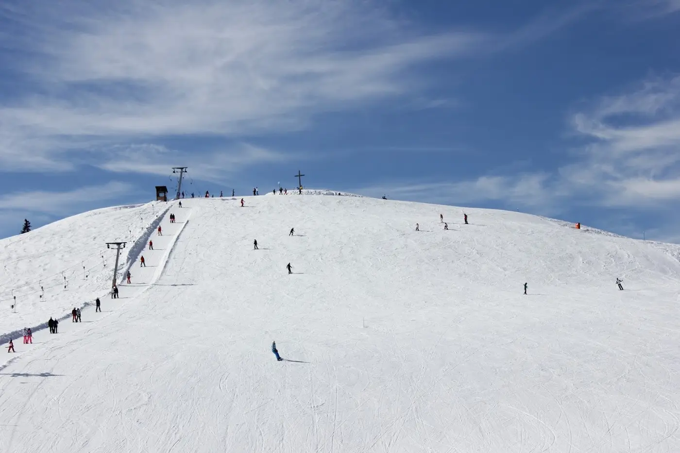 Wide snowy ski hill with a drag lift, skiers on the slope and a summit cross on top under a blue sky