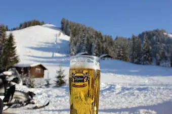 Glass of beer on a winter terrace with a snowy slope, a mountain hut and a snowmobile in the background