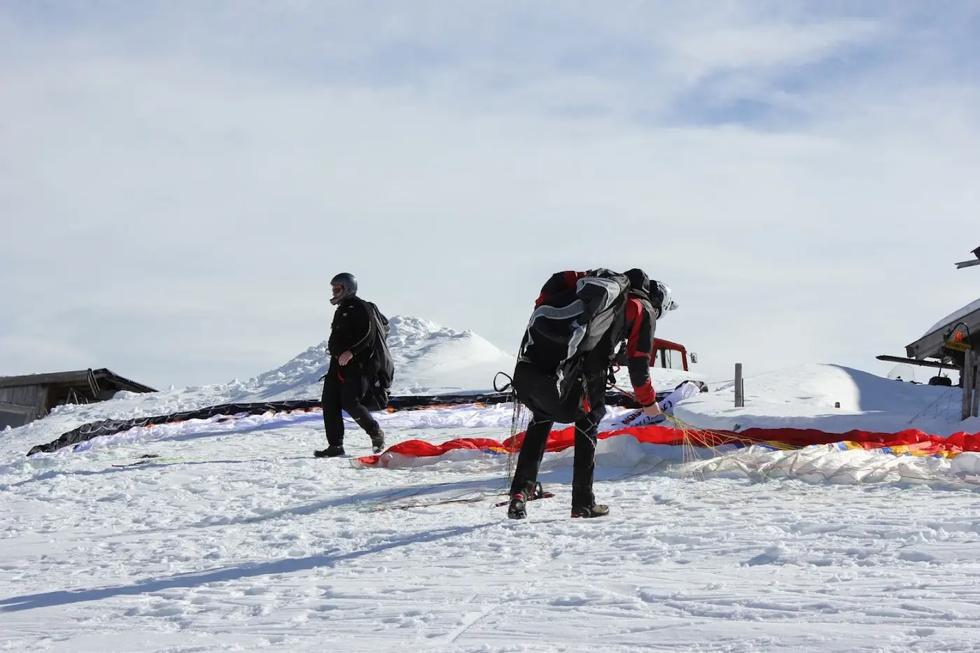 Two paragliders preparing their wings at a snowy take-off spot near a mountain hut