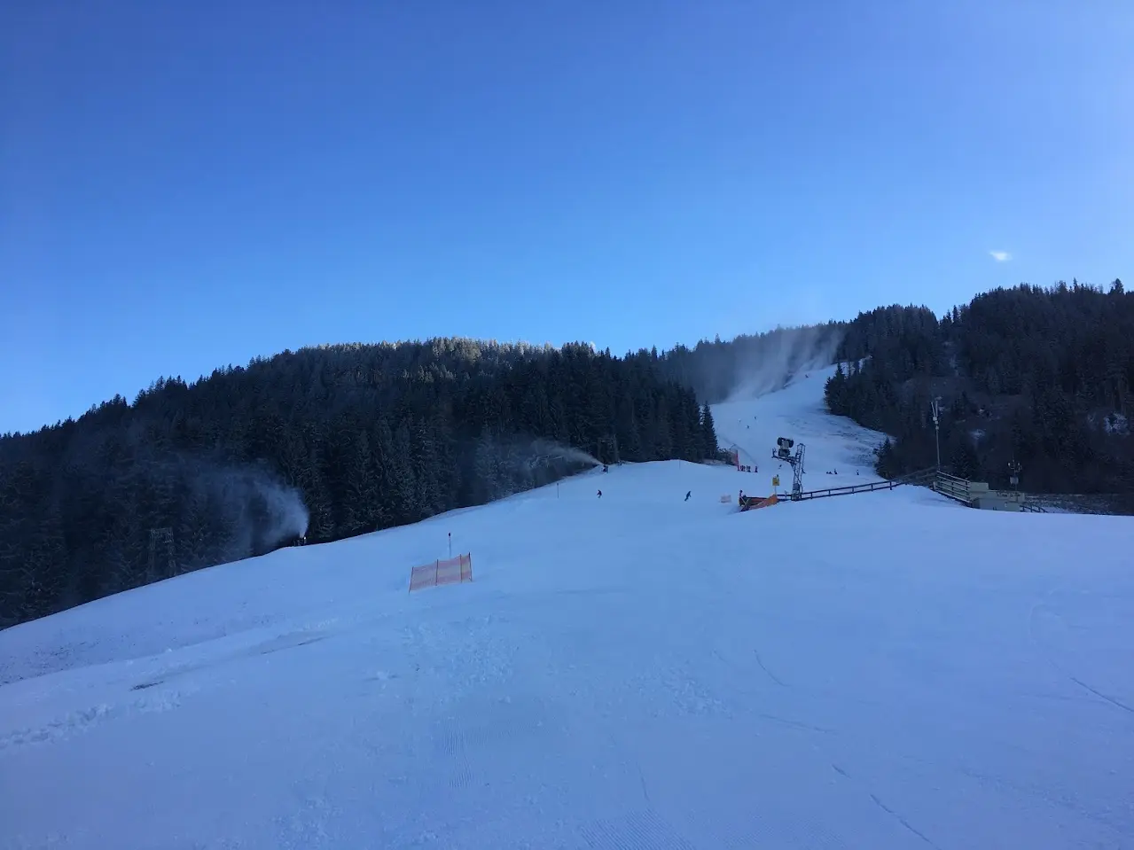 Ski slope with working snow cannons under a deep blue sky, with forest and a lift station