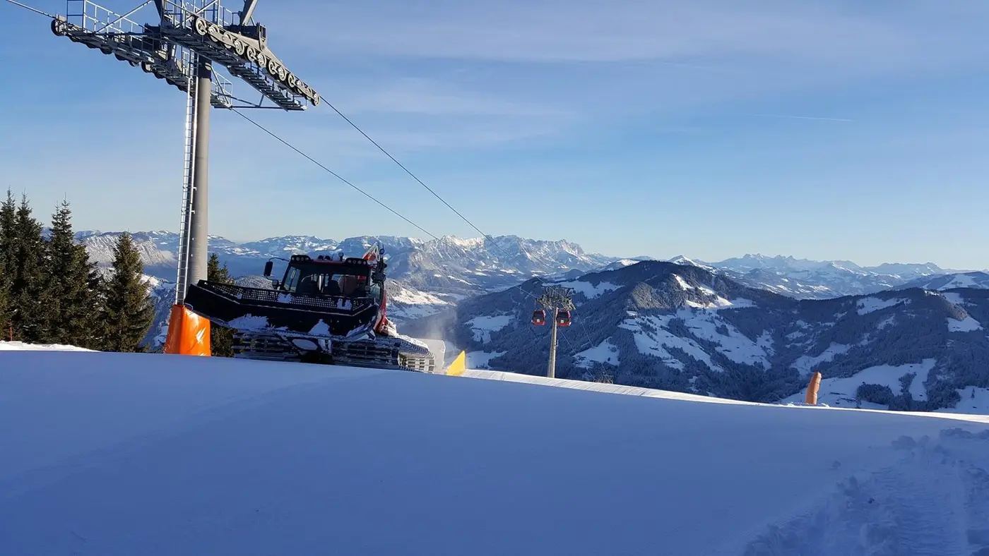 Snow groomer on a mountain top next to a gondola pylon, with a view over snowy mountain ranges