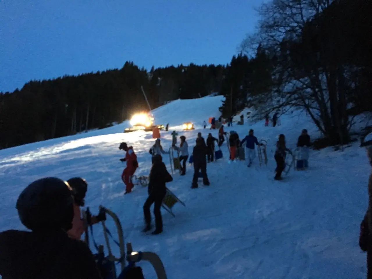 Tobogganers at dusk on a snowy slope, with a lit snow groomer in the background