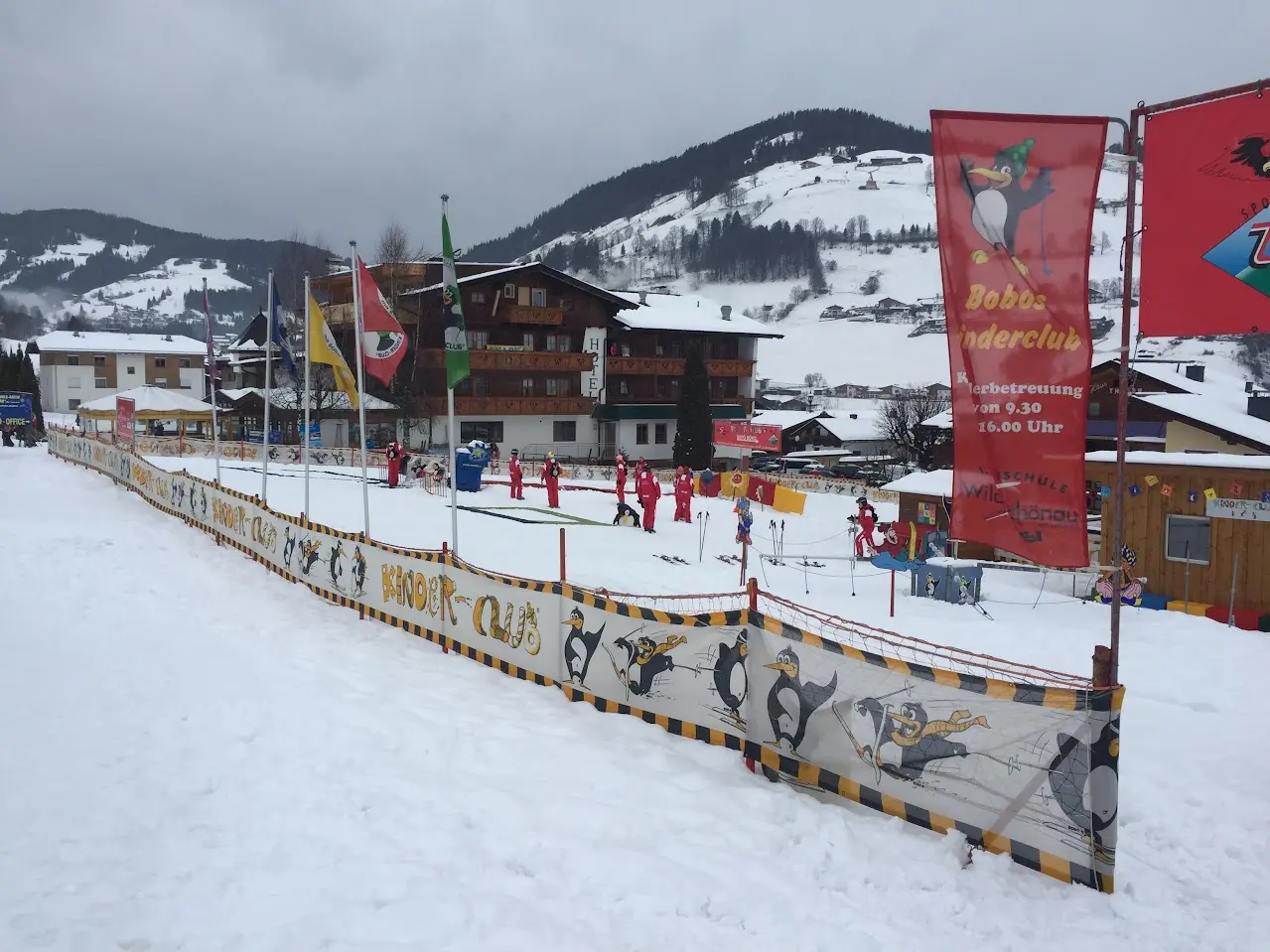École de ski pour enfants avec des banderoles rouges et une zone d’entraînement clôturée dans la neige, avec un village de montagne derrière