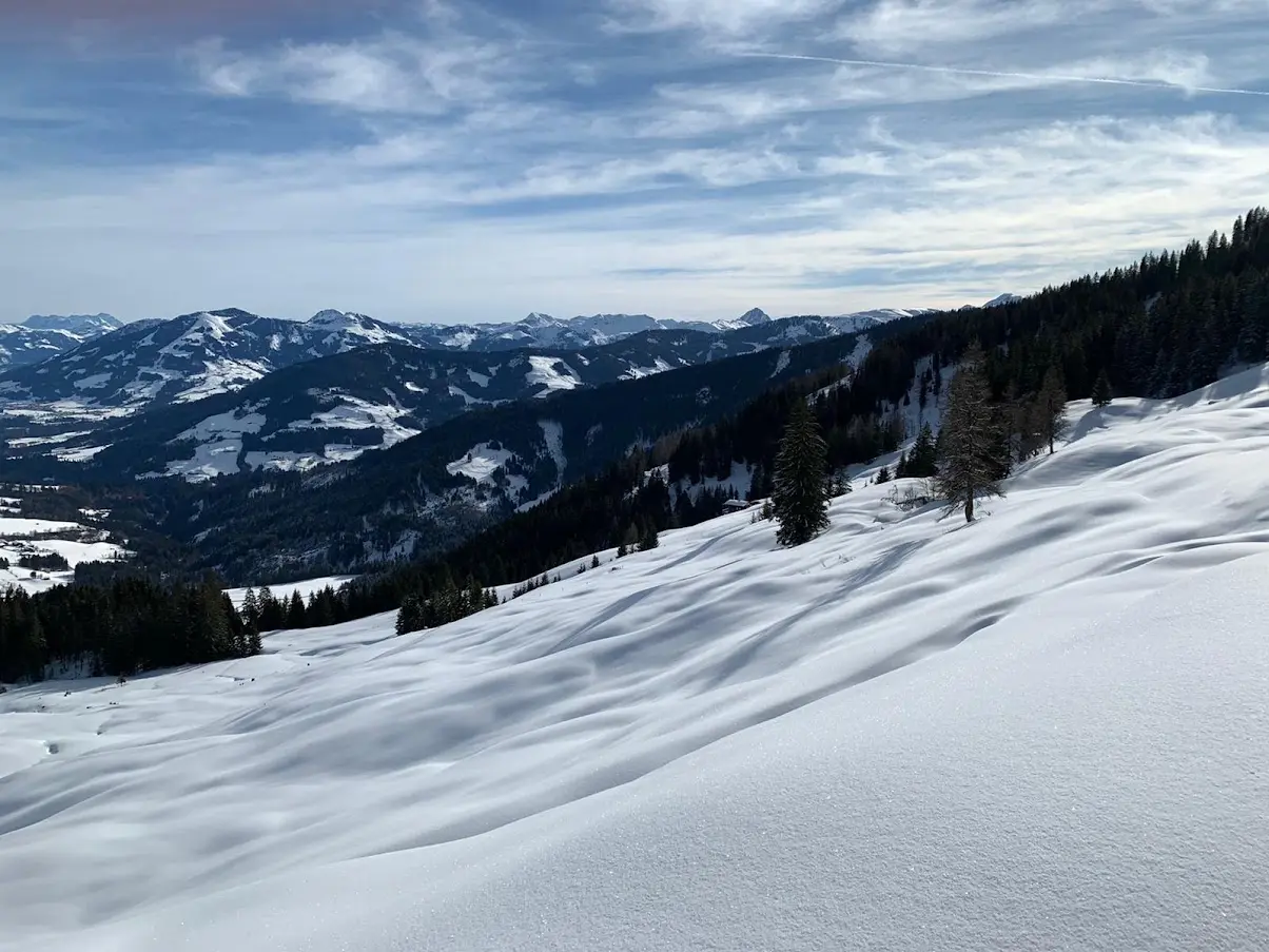 Snowy mountain slope with soft shadows and a view of mountain ranges under a streaky sky