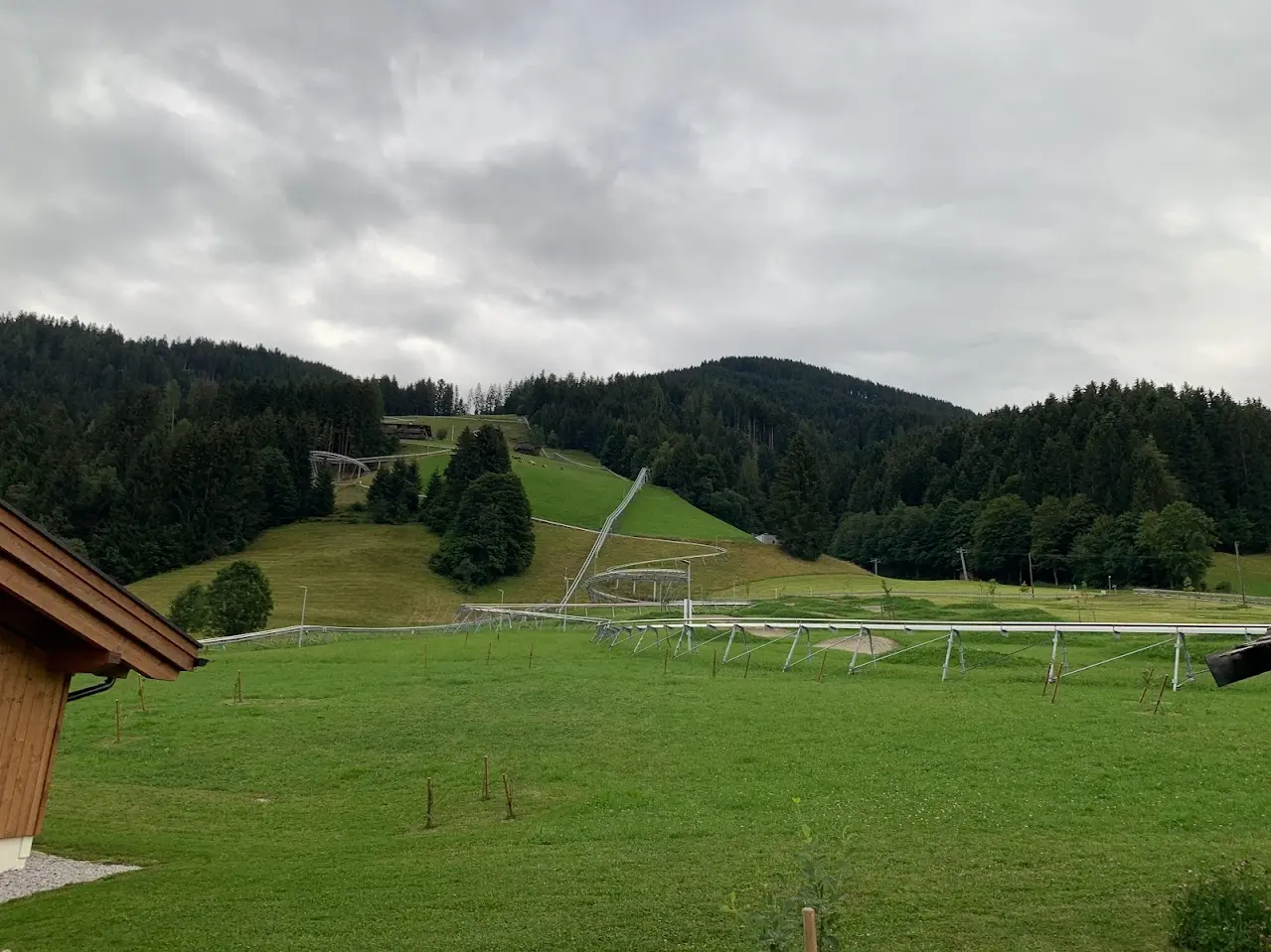 Steel track of an alpine coaster winding across a green slope under a grey sky