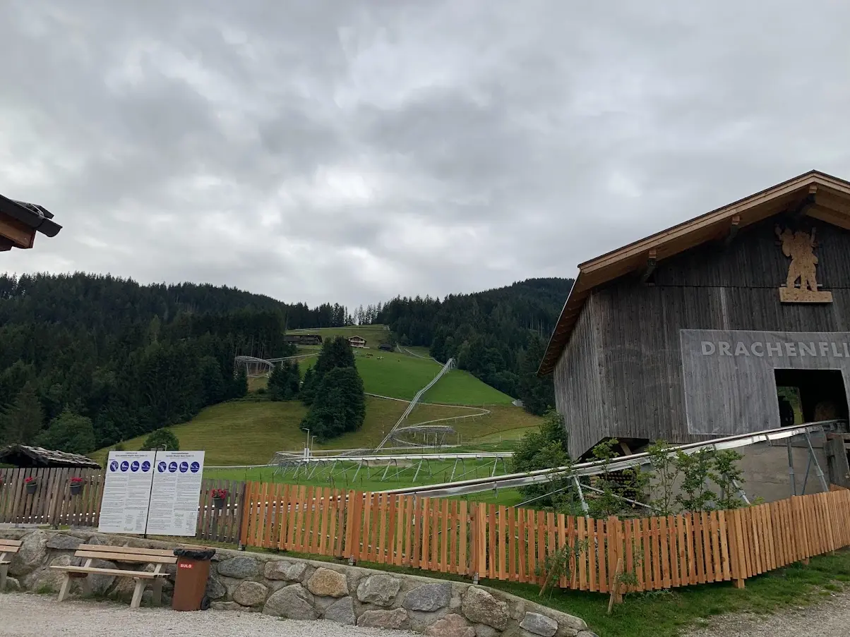 Wooden barn with a carved figure and the letters ‘Drachenfl…’, beside the rails of an alpine coaster on a green slope