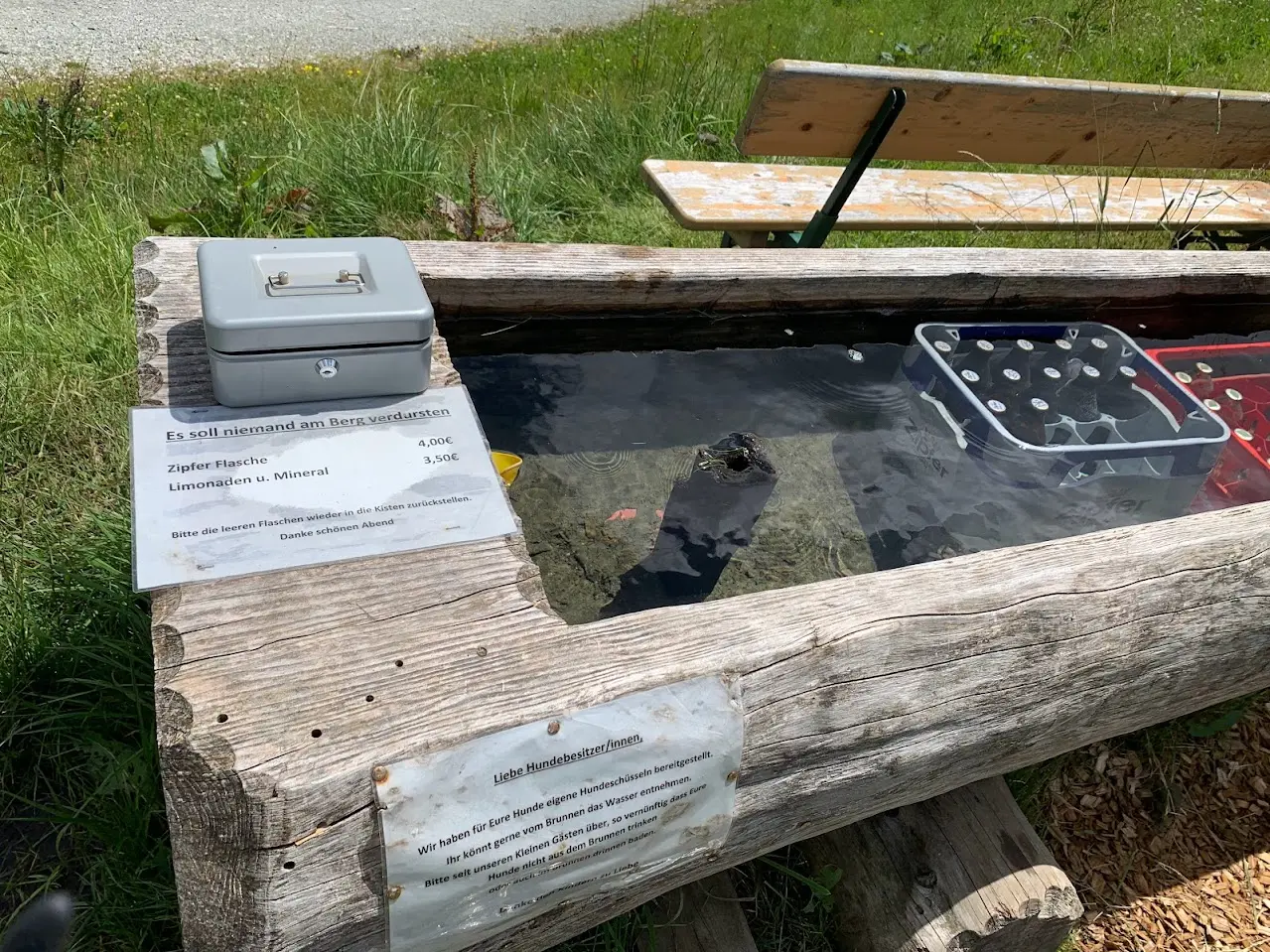 Wooden trough of mountain water cooling crates of bottles, with a cash box and a sign with drink prices and a notice for dog owners