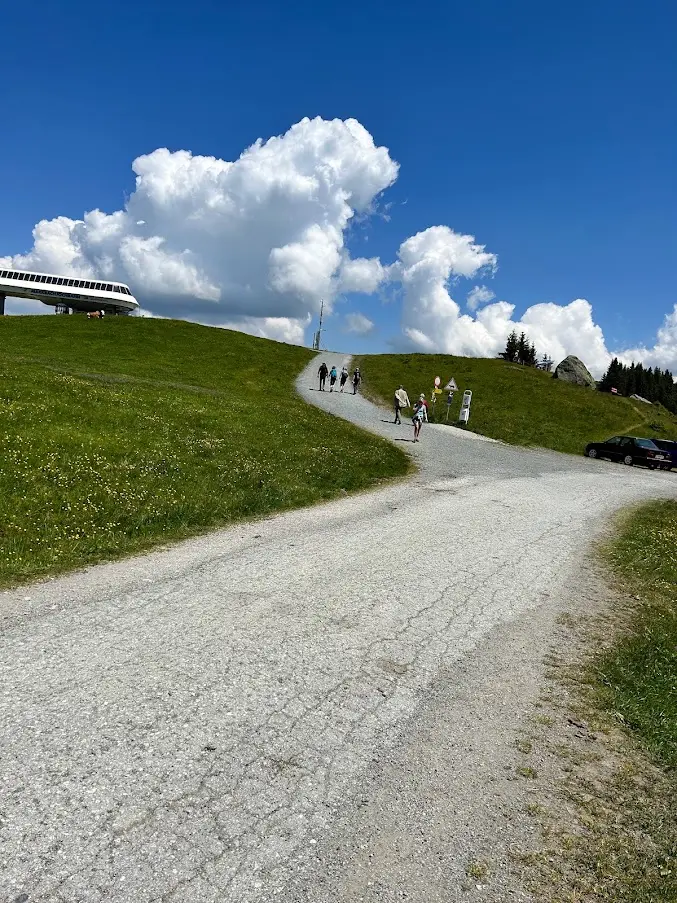 Gravel path across a grassy slope towards a mountain station and walkers, under a blue sky with big clouds