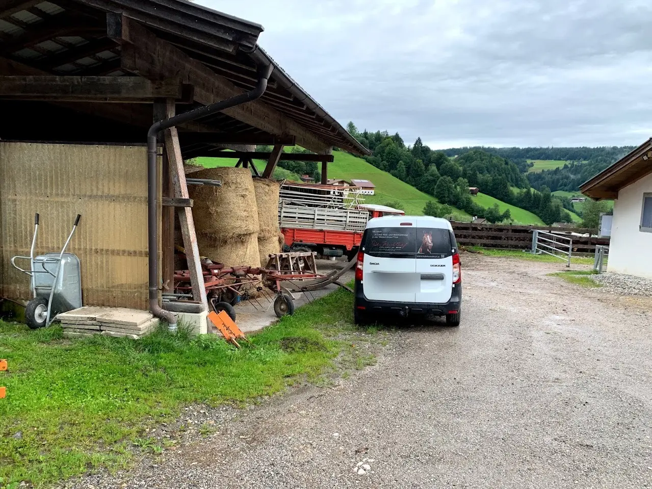 Farmyard on a rainy day with hay bales under a shelter, a wheelbarrow, a hay tedder and a white van, with green hills behind