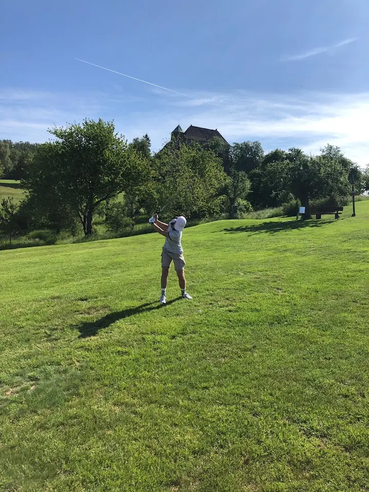 Golfer in the follow-through of a shot on a hilly fairway, with a castle-like building among the trees