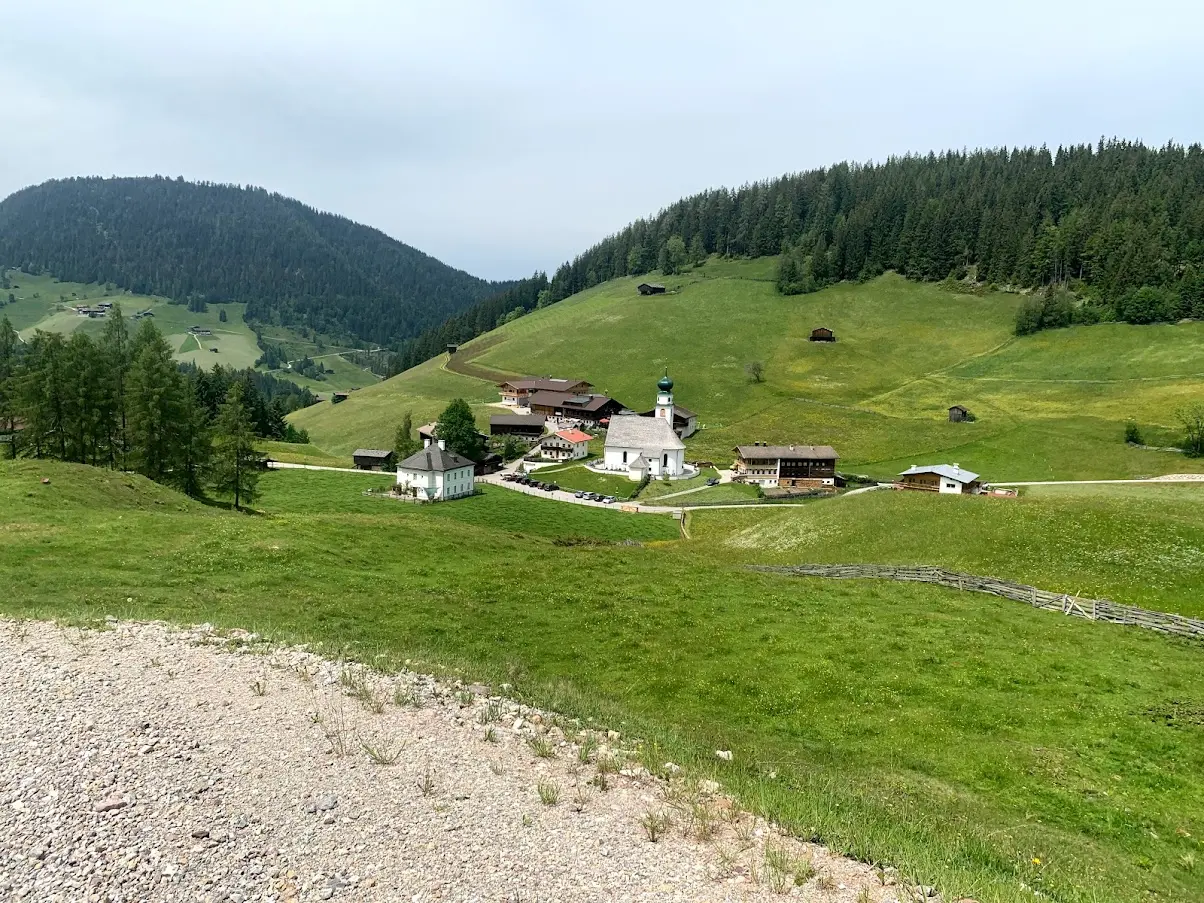 White chapel with an onion-shaped spire and a few farmhouses in a hamlet among green meadows and wooded hills