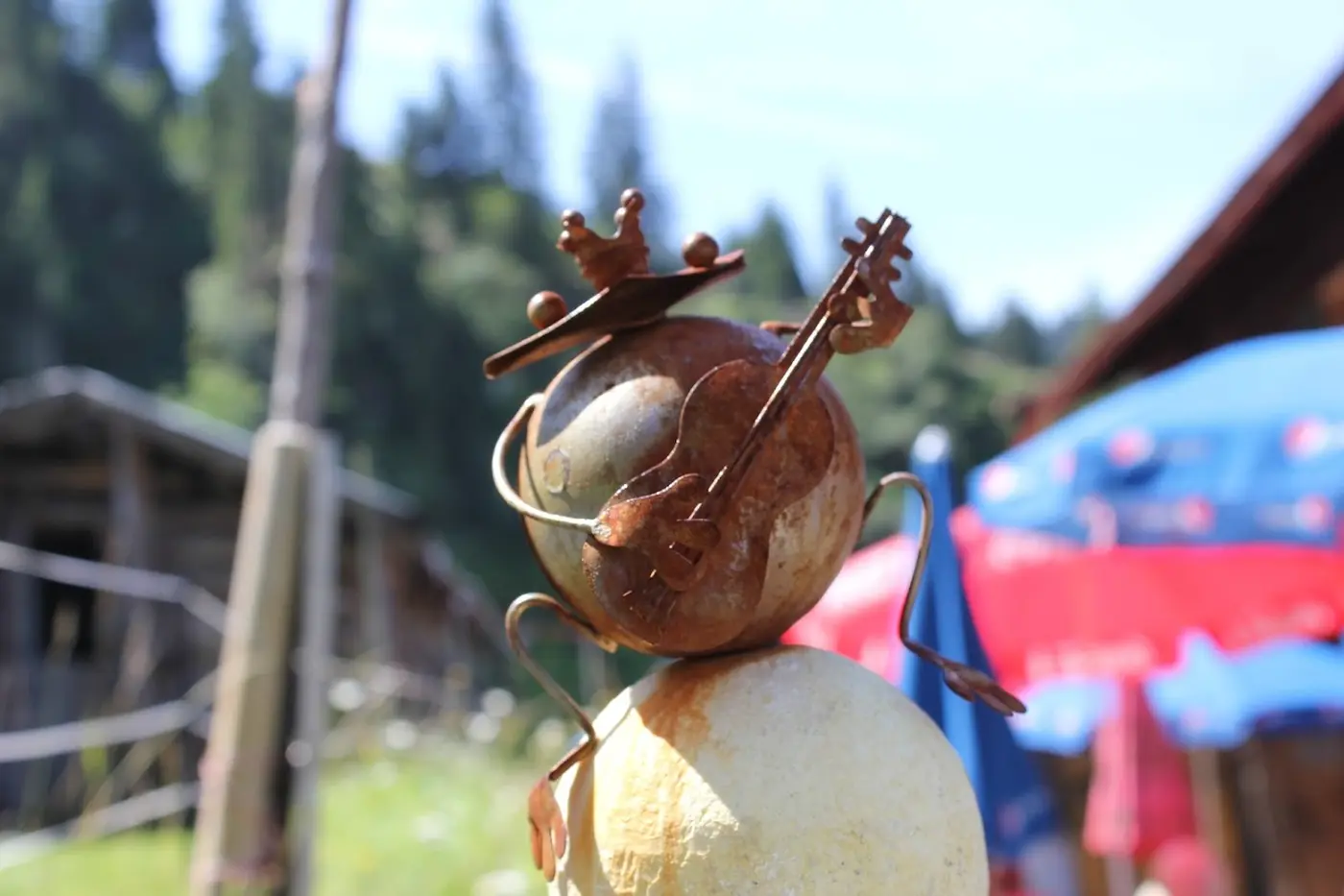 Rusty metal artwork of a frog king with a crown and guitar at a mountain hut, with parasols in the background