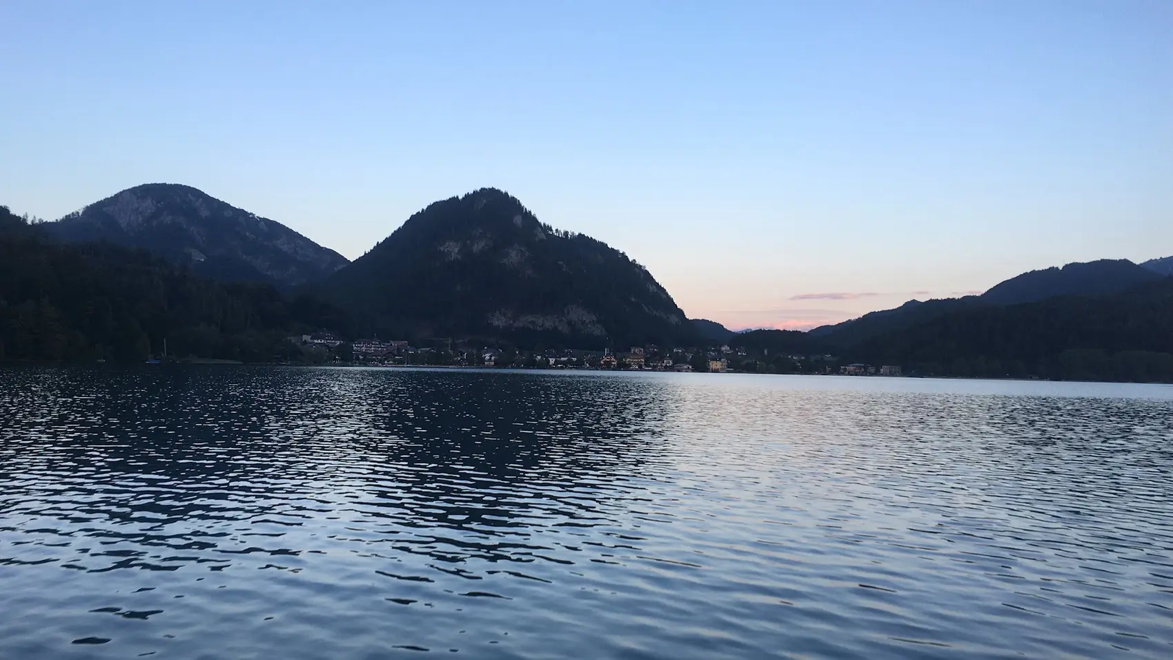 Mountain lake at dusk with dark mountains and a village on the shore under a pink-blue sky