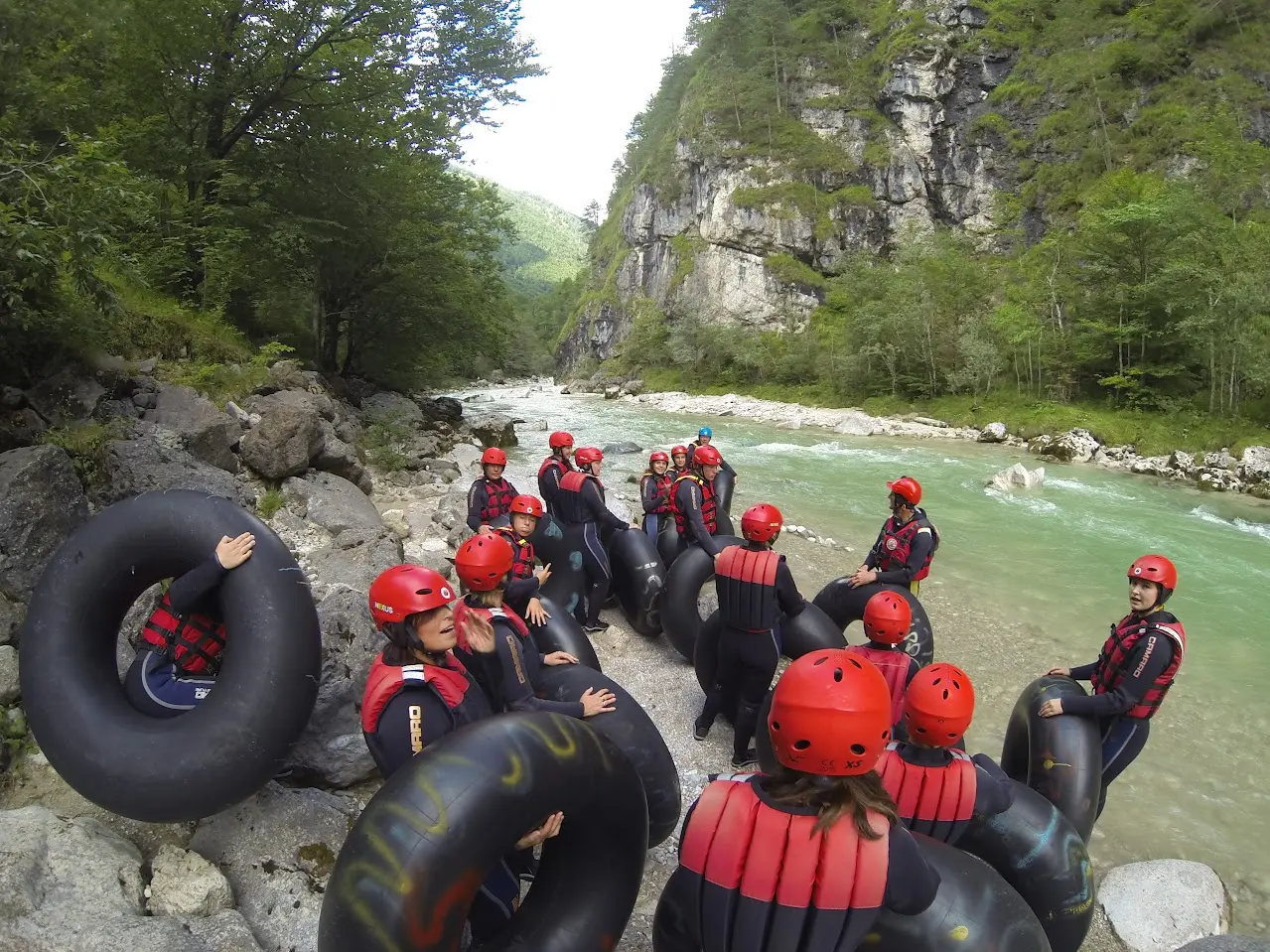Group in wetsuits, red helmets and life vests with black inner tubes on the bank of a green mountain river, by a rock face