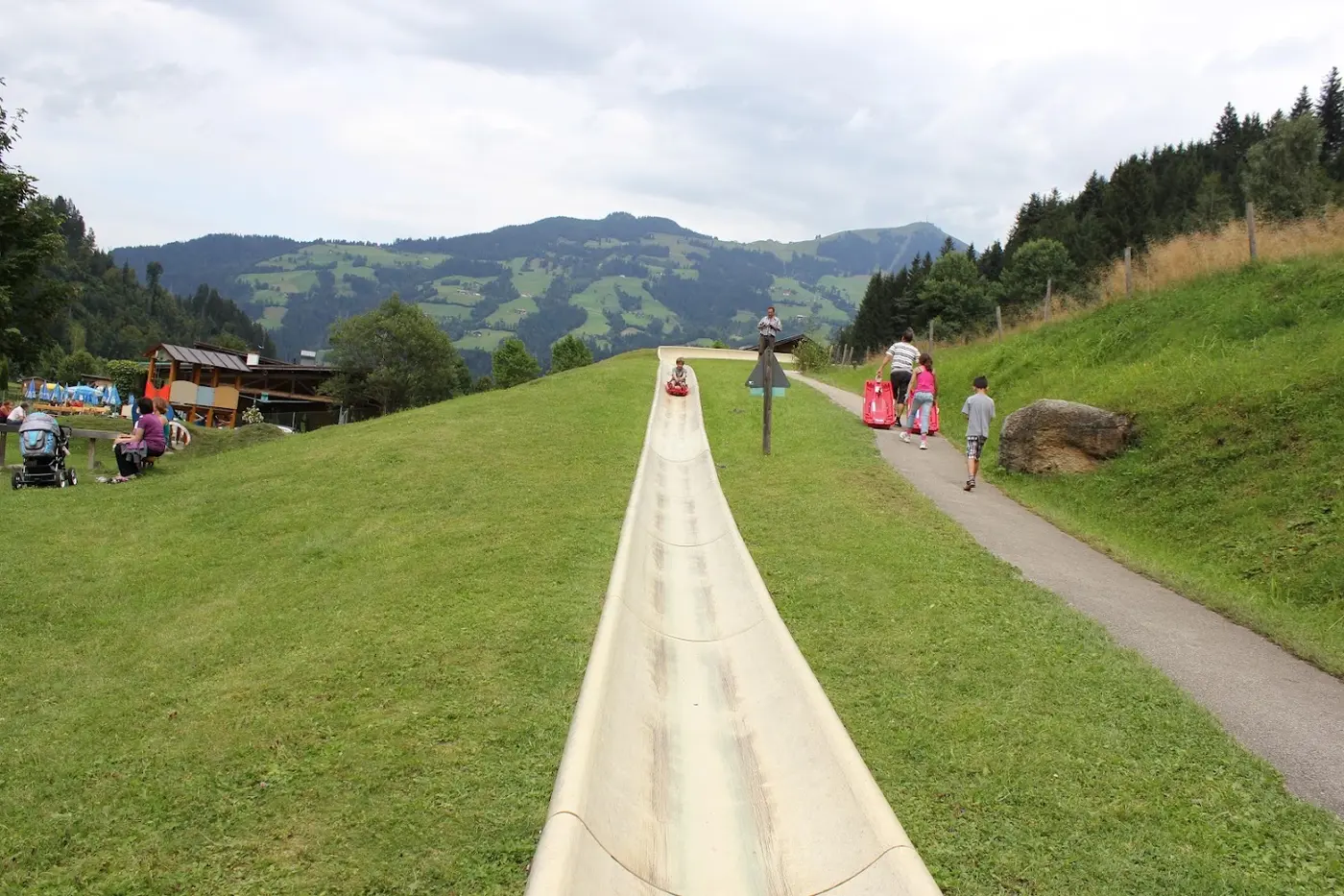 Summer toboggan run: a child slides in a sled down a long chute across a grassy slope, with mountains behind