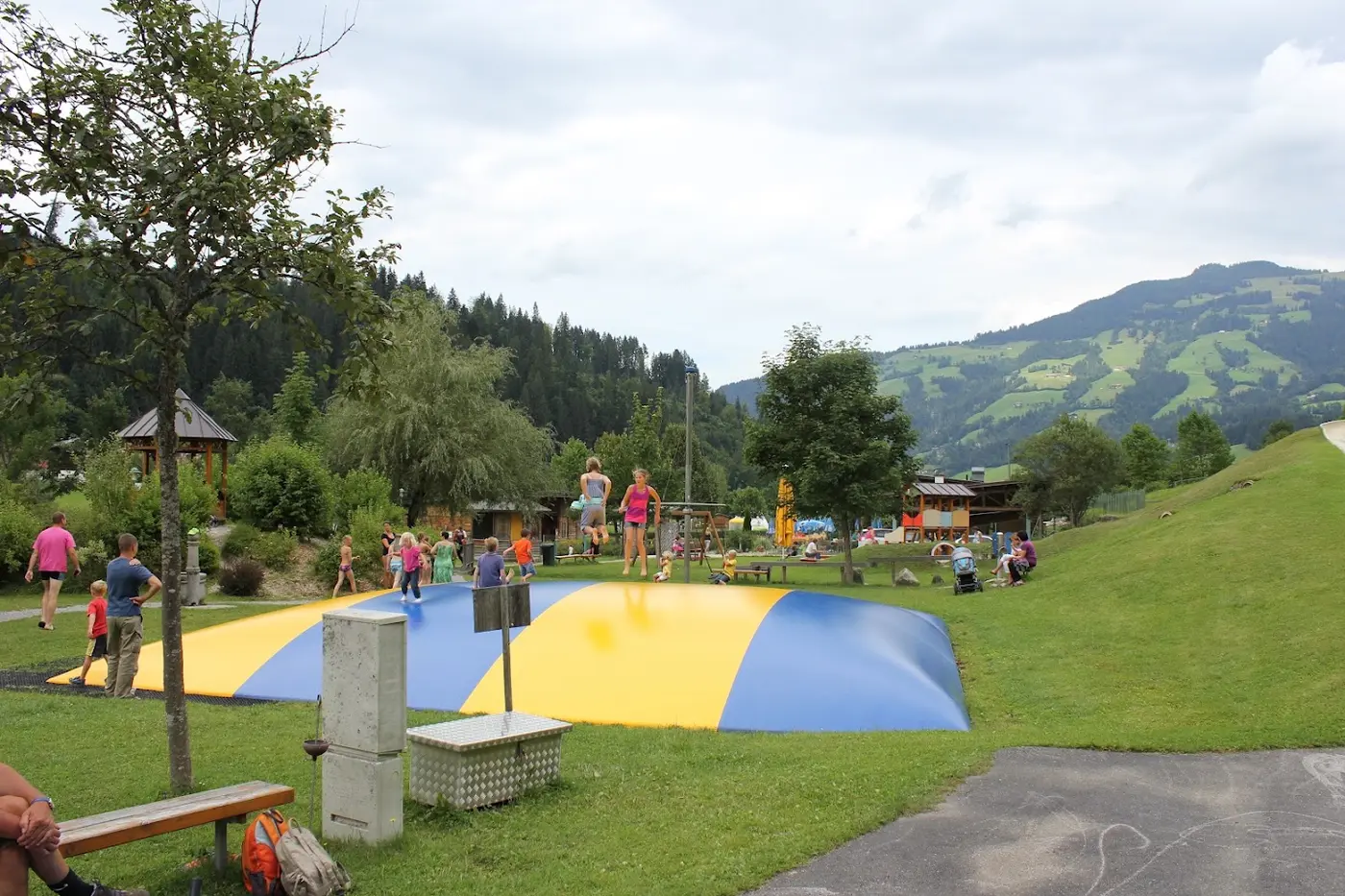 Children jumping on a large yellow and blue jumping pillow in a grassy playground, with mountains behind