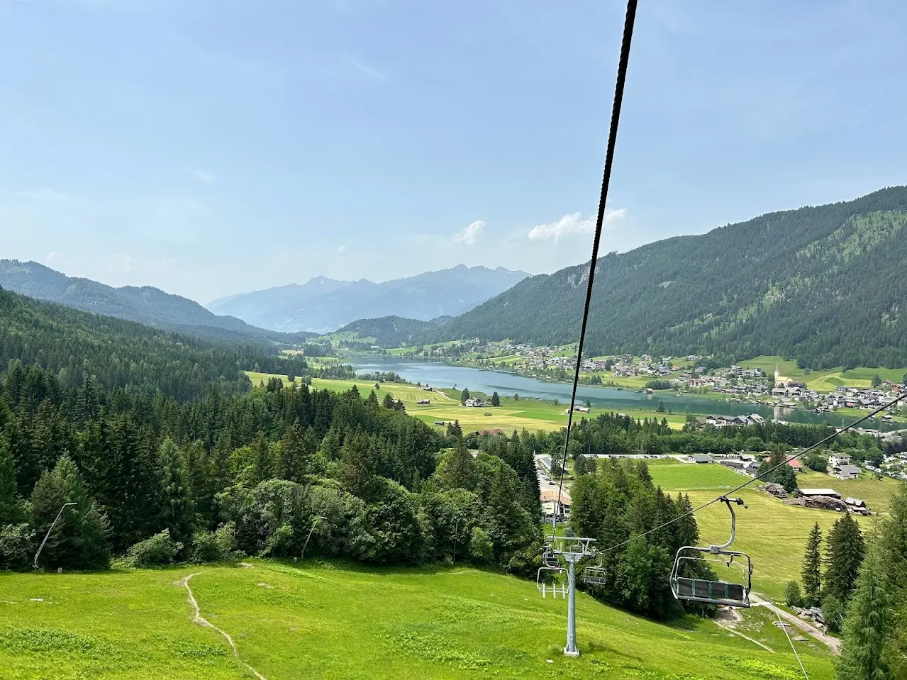 View from a chairlift onto a mountain lake, a village with a church and wooded mountainsides in summer