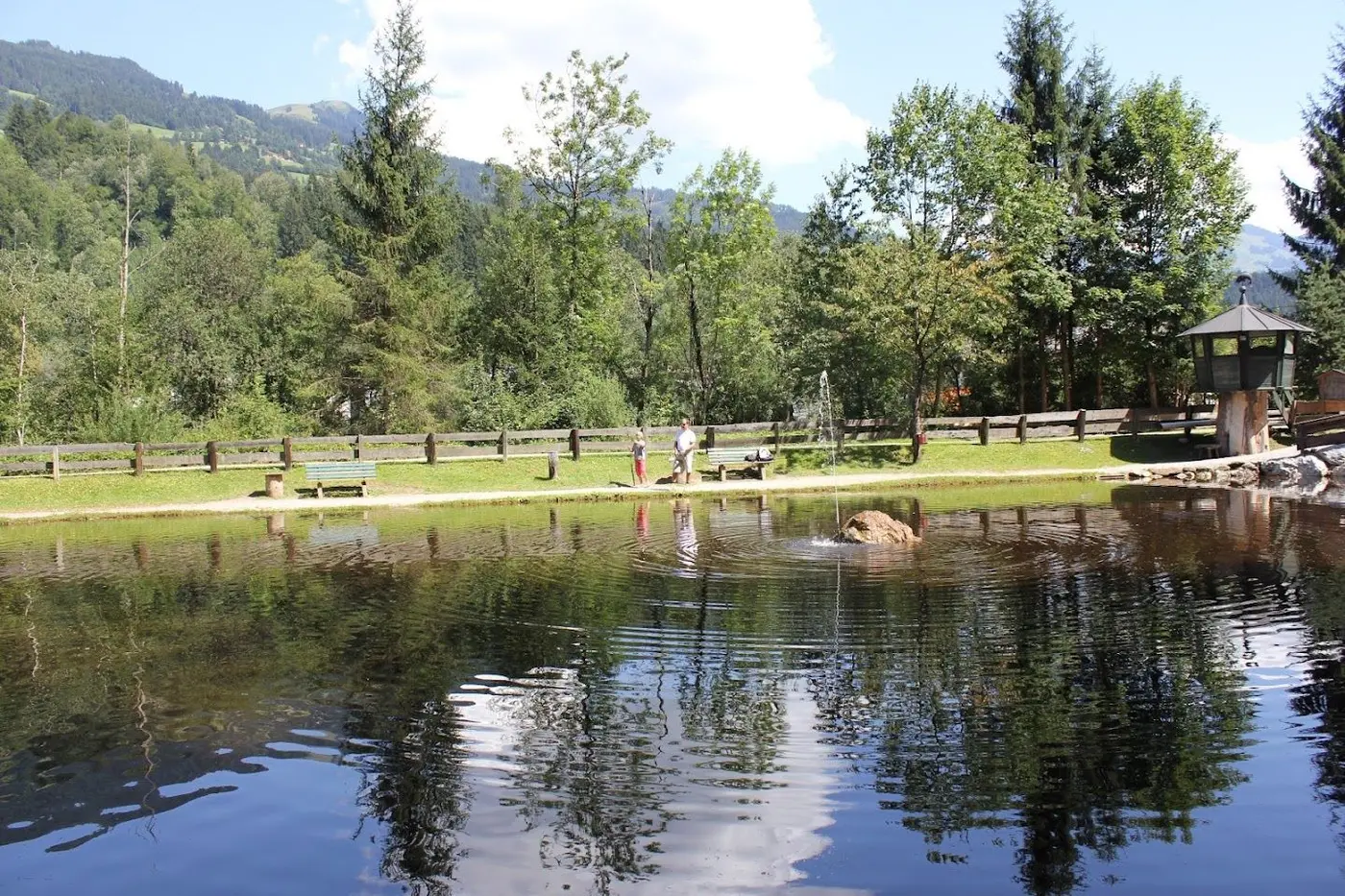 Pond with a small fountain, benches and a lookout hut among trees, with mountains in the background