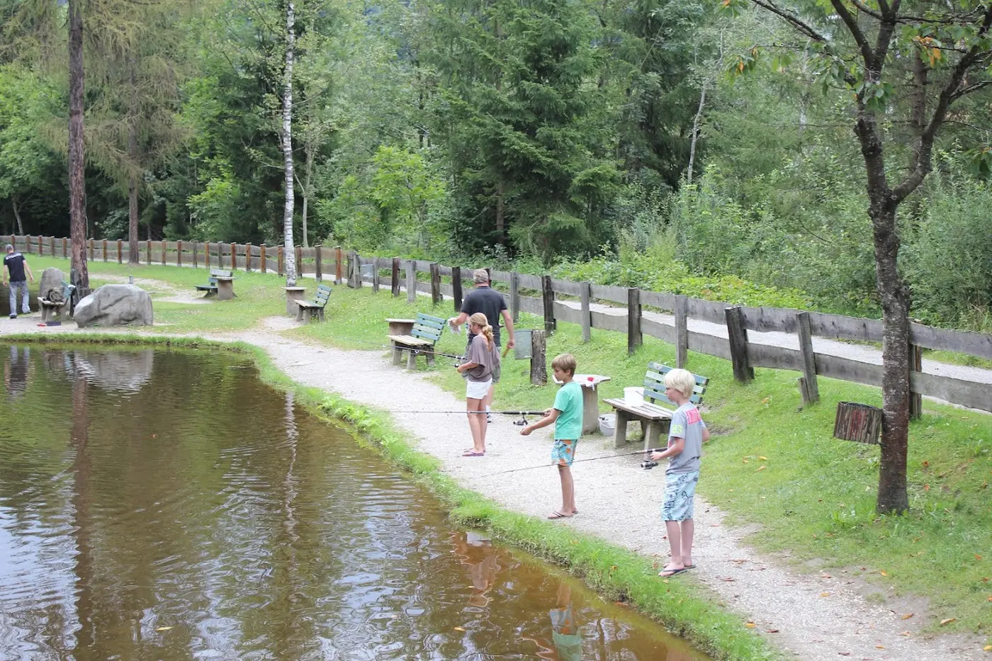 Children and an adult fishing at a pond surrounded by forest and a gravel path