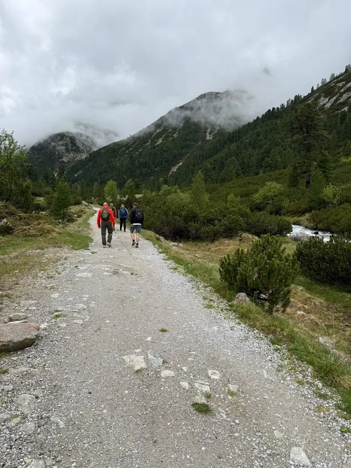 Hikers on a wide gravel track in a mountain valley with a stream and clouds hanging around the peaks