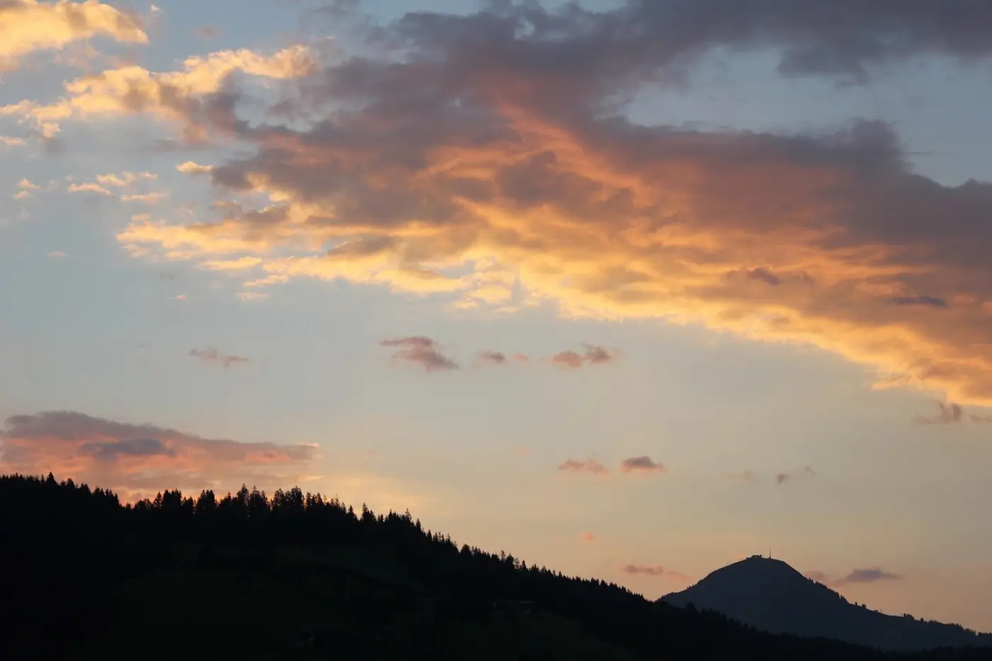 Sunset with orange clouds above a wooded ridge and the silhouette of a mountain with a mast on its summit